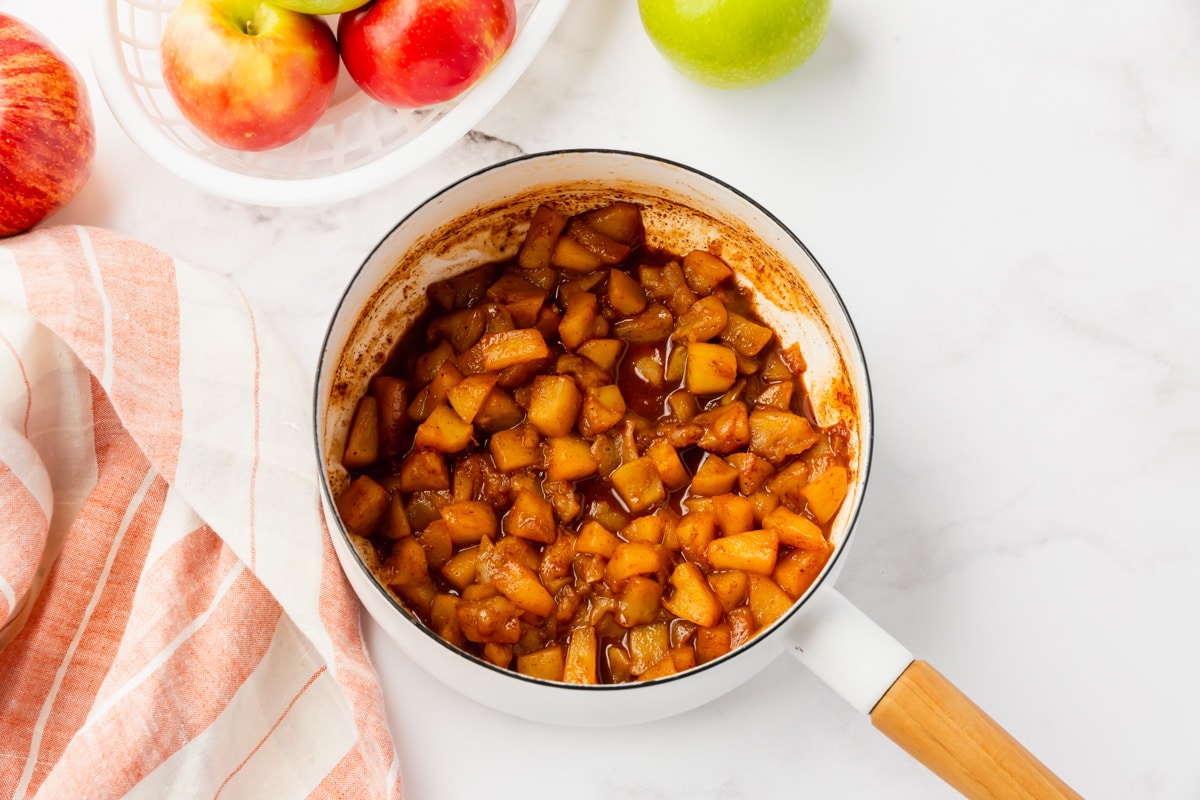 cooked brown apples in sauce in a white sauce pan with napkin to the left and apples to the top left