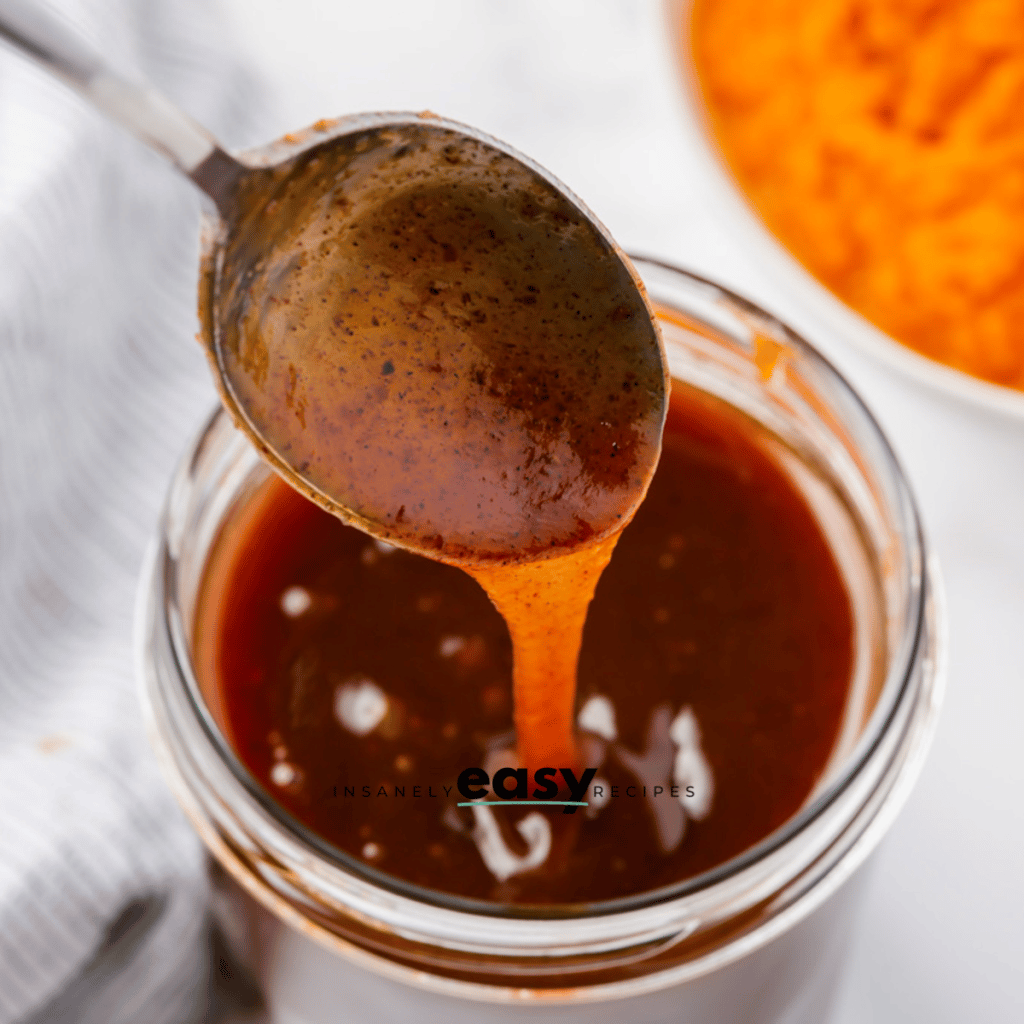 glass jar with brown liquid, metal spoon dipping into the liquid and some pouring off spoon. Top right corner has puree pumpkin in a white bowl. Gray napkin to left side.