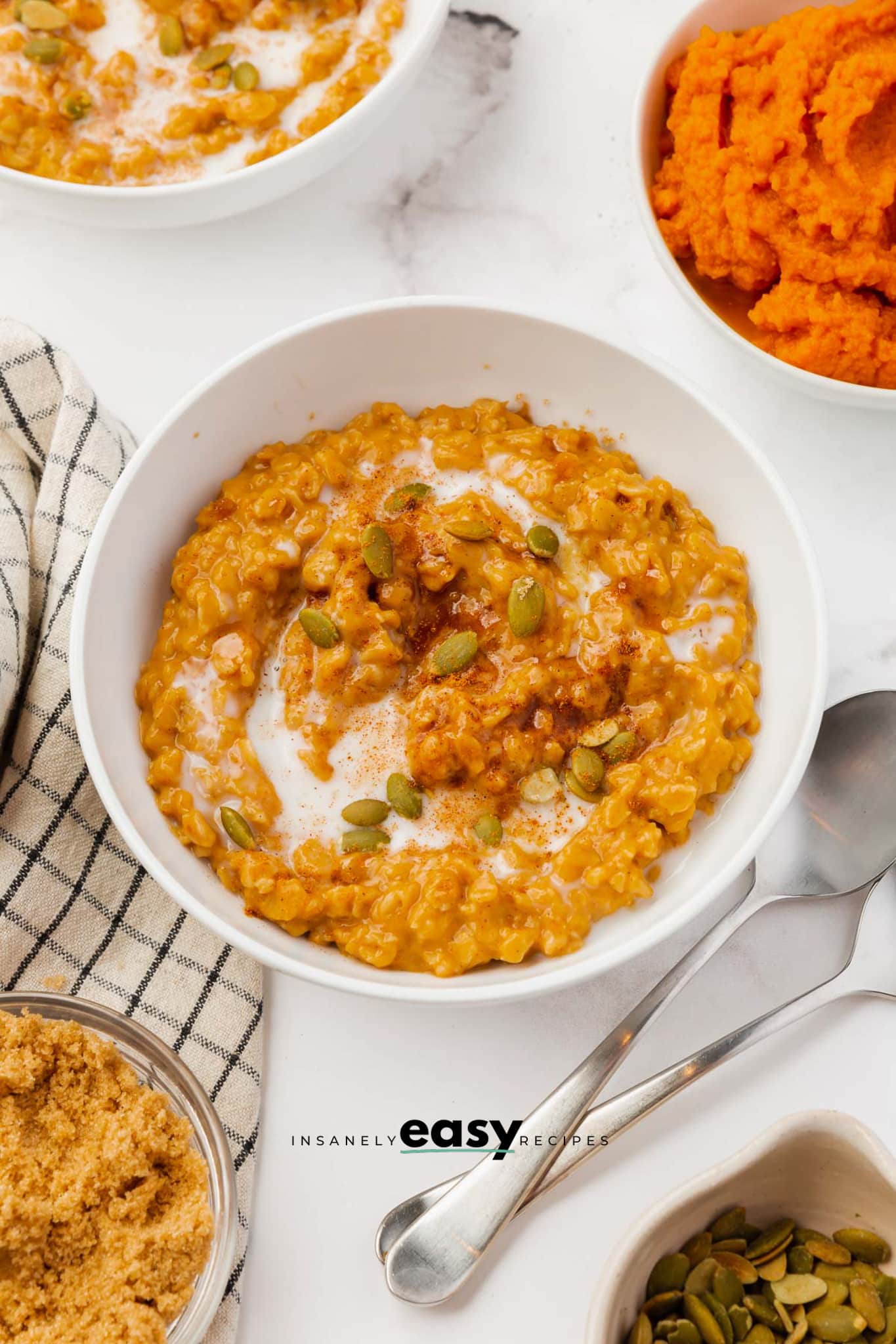 A white bowl filled with pumpkin oatmeal, topped with pepitas. Around the bowl is other bowls of the ingredients used to make the oatmeal.