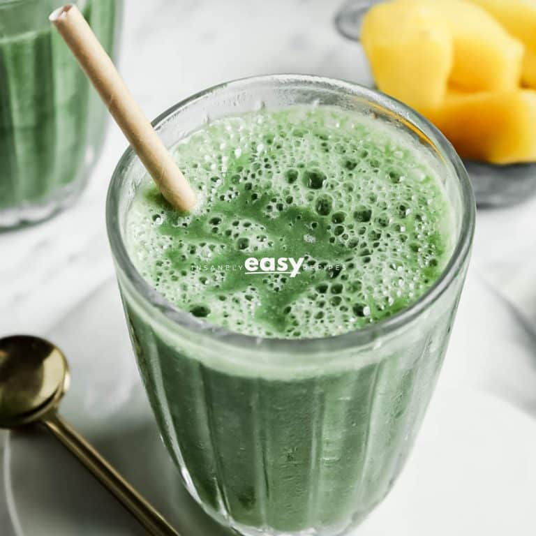 Closeup photo of Spirulina Smoothie in a glass jar with a paper straw, on a white plate with a gold spoon. There are frozen pineapples in the background and another glass of smoothie.