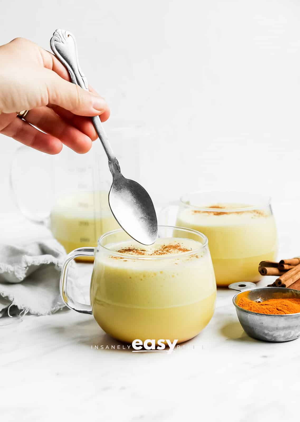 Photo of Turmeric Coffee in a glass mug. There is a hand holding a silver spoon, ready to dip into the mug. There is powdered turmeric to the right of the glass and cinnamon sticks. There is a gray kitchen towel to the left of the glass. And there is a mug with turmeric coffee in the background. 