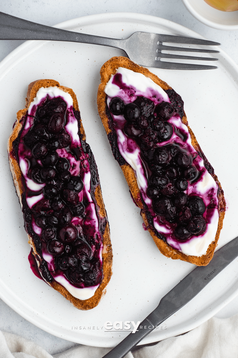 top view photo of 2 slices of blueberry toast on a white plate with a fork and small bowl of honey above it, and a butter knife and white kitchen towel below it