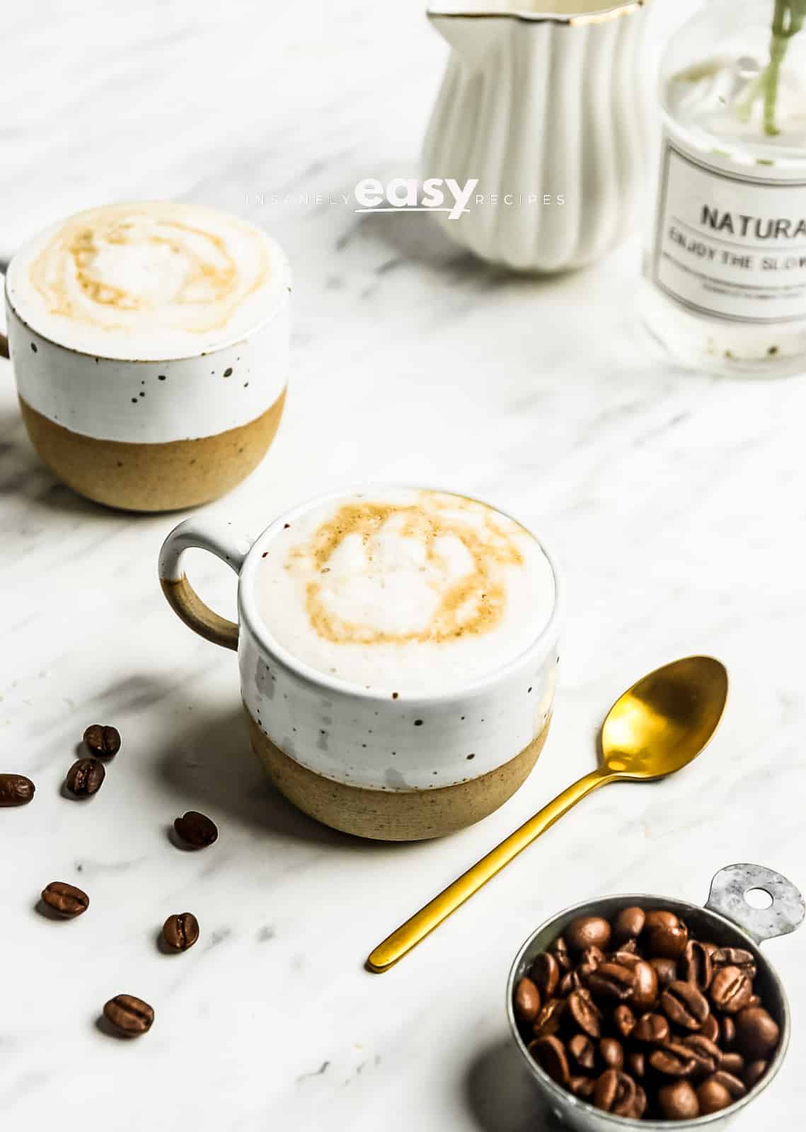 Photo of two mugs filled with Oat milk Latte. There is a gold spoon next to the mug, a measuring cup filled with coffee beans, and a small pitcher with oat milk in the background. 