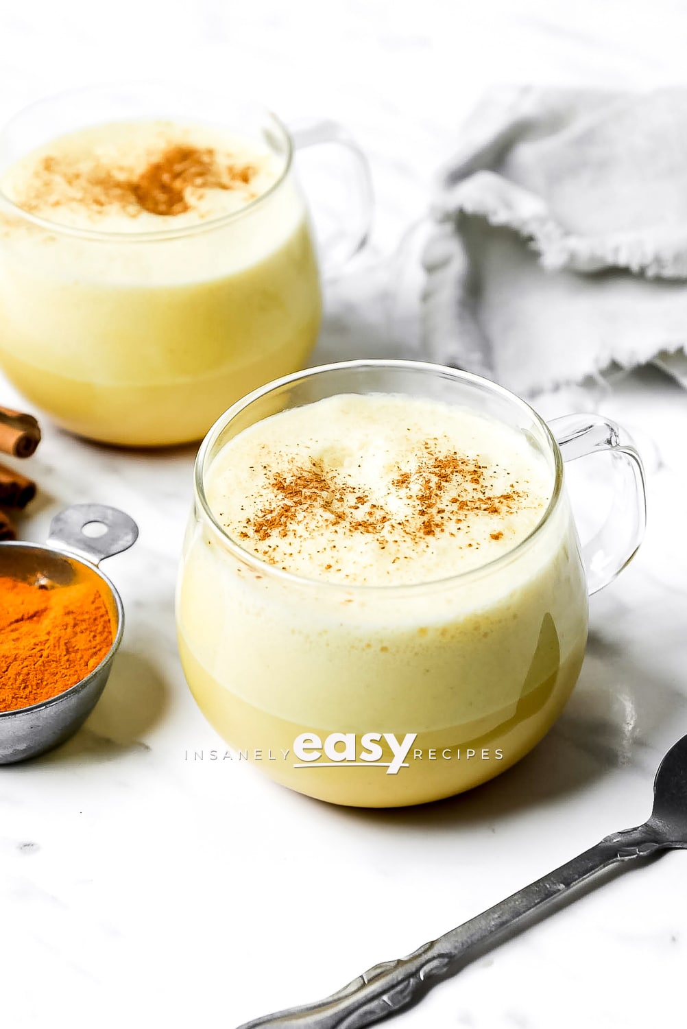 Photo of two glass mugs with Turmeric coffee in them, with a silver spoon in the foreground, turmeric powder on the right side, and a gray kitchen towel in the background.