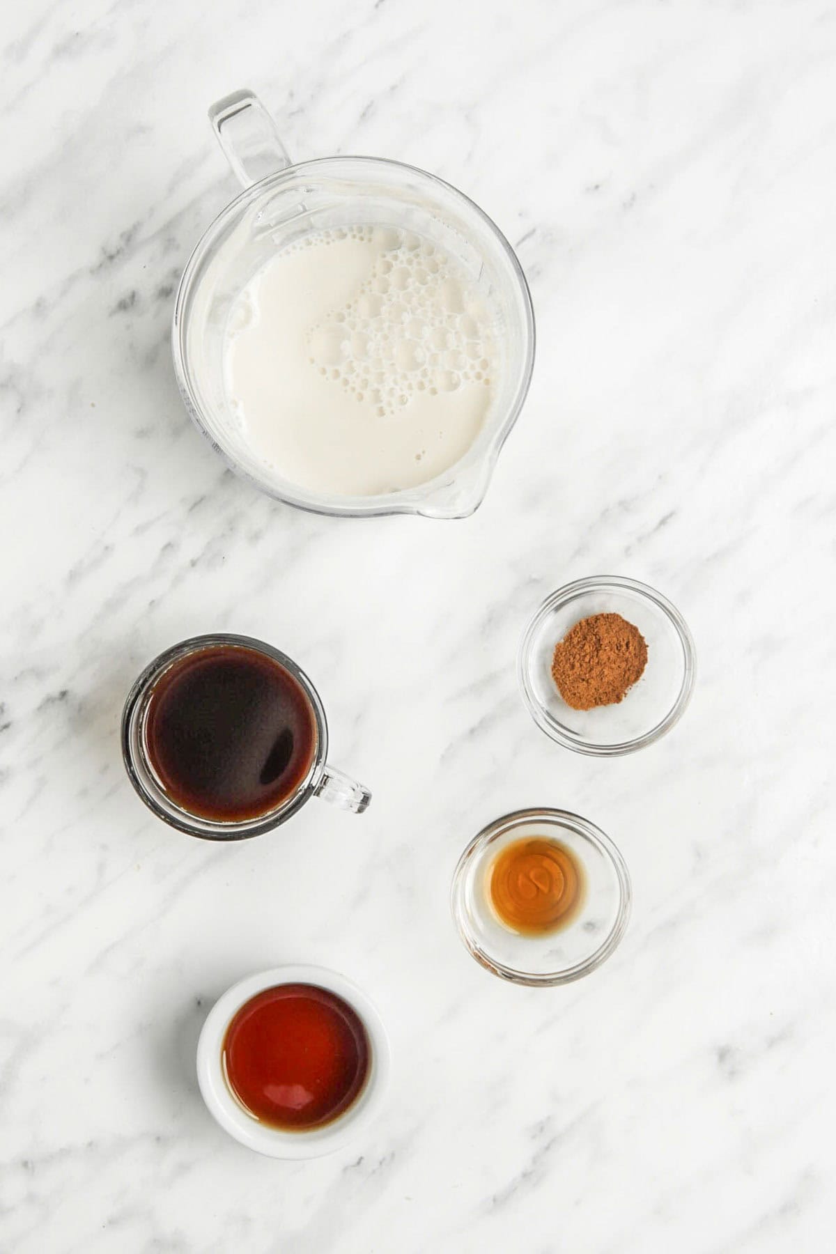 Five small containers on a marble surface, each holding an ingredient for an oat milk latte: milk, brewed coffee, cinnamon, vanilla extract, and maple syrup.