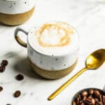 A cup of cappuccino with foam and caramel drizzle sits on a marble surface next to a gold spoon, coffee beans, and an oat milk latte in the background.