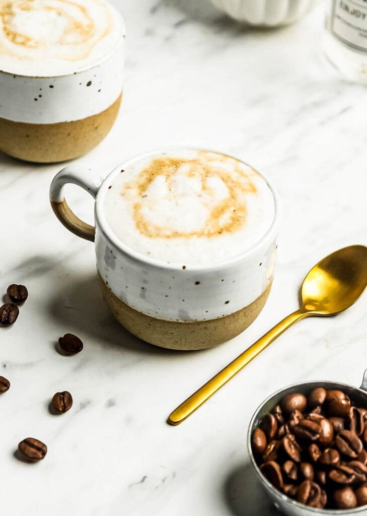 A cup of cappuccino with foam and caramel drizzle sits on a marble surface next to a gold spoon, coffee beans, and an oat milk latte in the background.