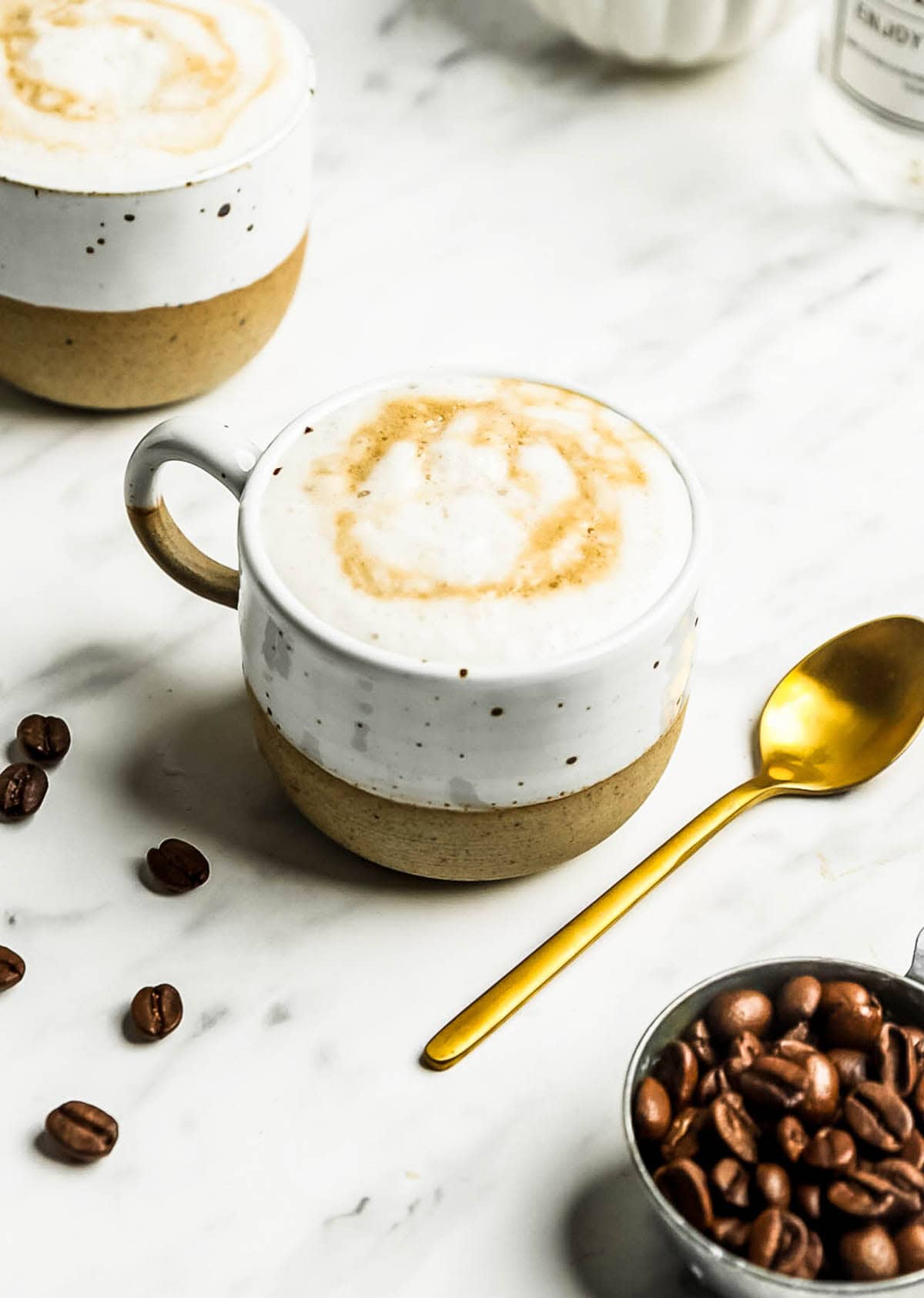 A cup of cappuccino with foam and caramel drizzle sits on a marble surface next to a gold spoon, coffee beans, and an oat milk latte in the background.