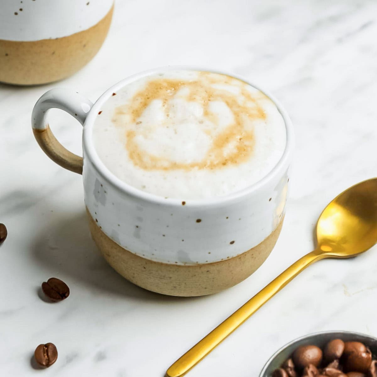 A ceramic mug filled with a frothy oat milk latte sits on a marble surface, next to a gold spoon, coffee beans, and a small container of chocolate chips.
