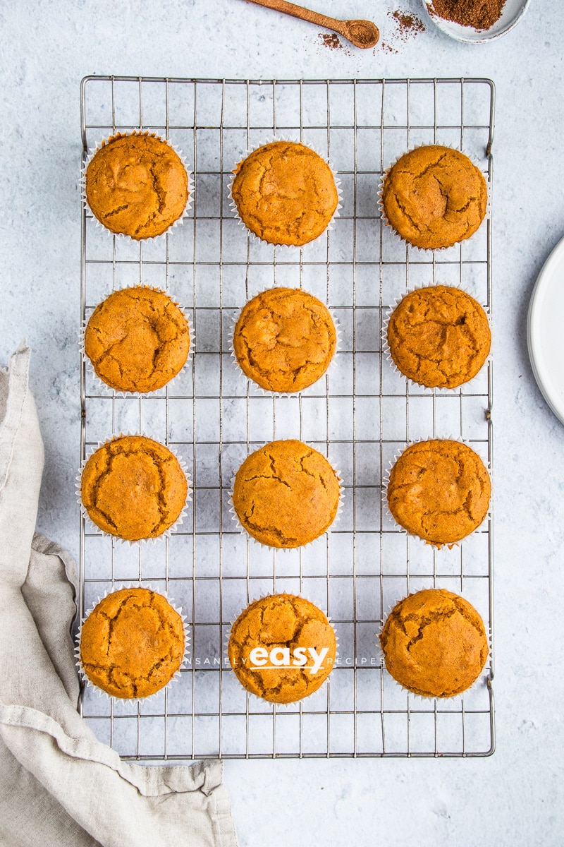 Topview photo of 12 Vegan Pumpkin Muffins, cooling on a wire rack. There is a kitchen towel to the left of the rack and pumpkin spice above the rack.