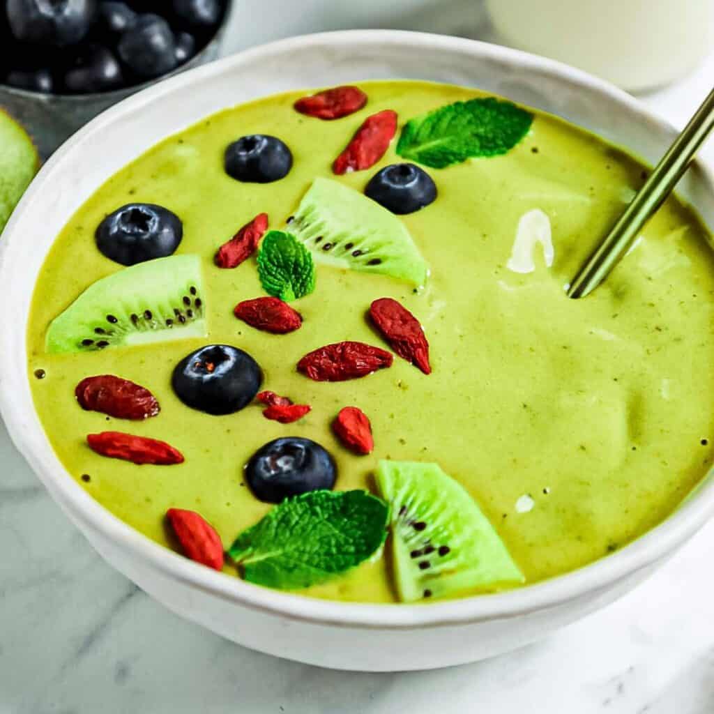 A creamy matcha bowl is topped with sliced kiwi, blueberries, goji berries, and mint leaves. A spoon rests in the bowl, with a small dish of blueberries visible in the background on a marble surface.