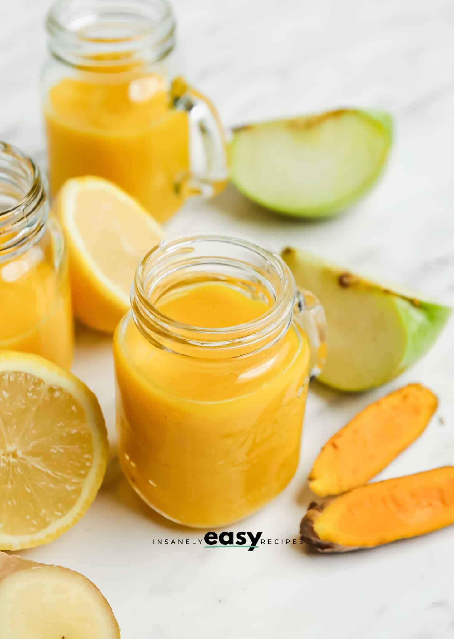 a small mason jar mug filled with orange ginger turmeric juice. Around the jar are pieces of apples, lemon, ginger, and turmeric root