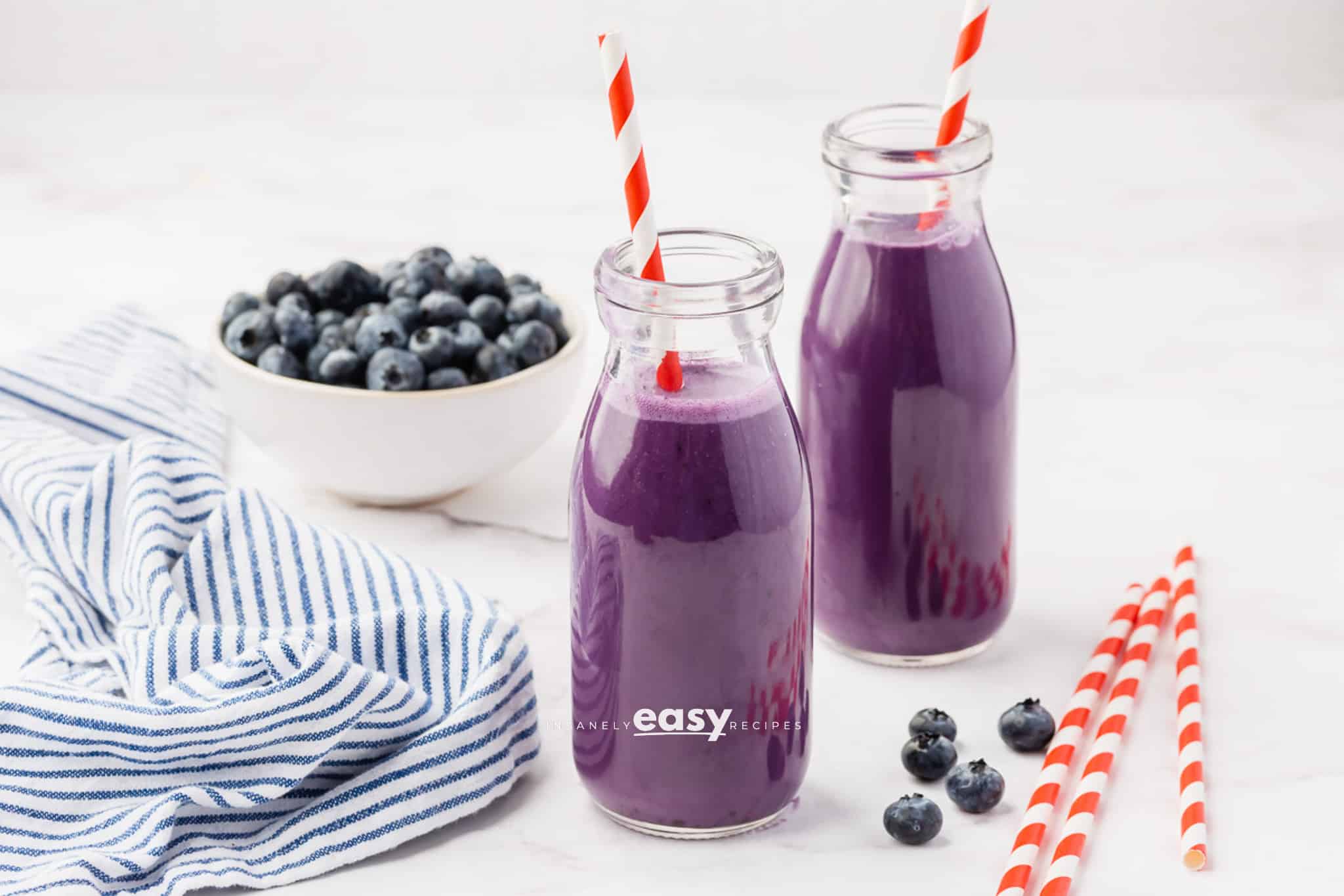 Two small glass jars filled with purple drink. There are red and white straws in each jar, and a bowl of blueberries in the background.