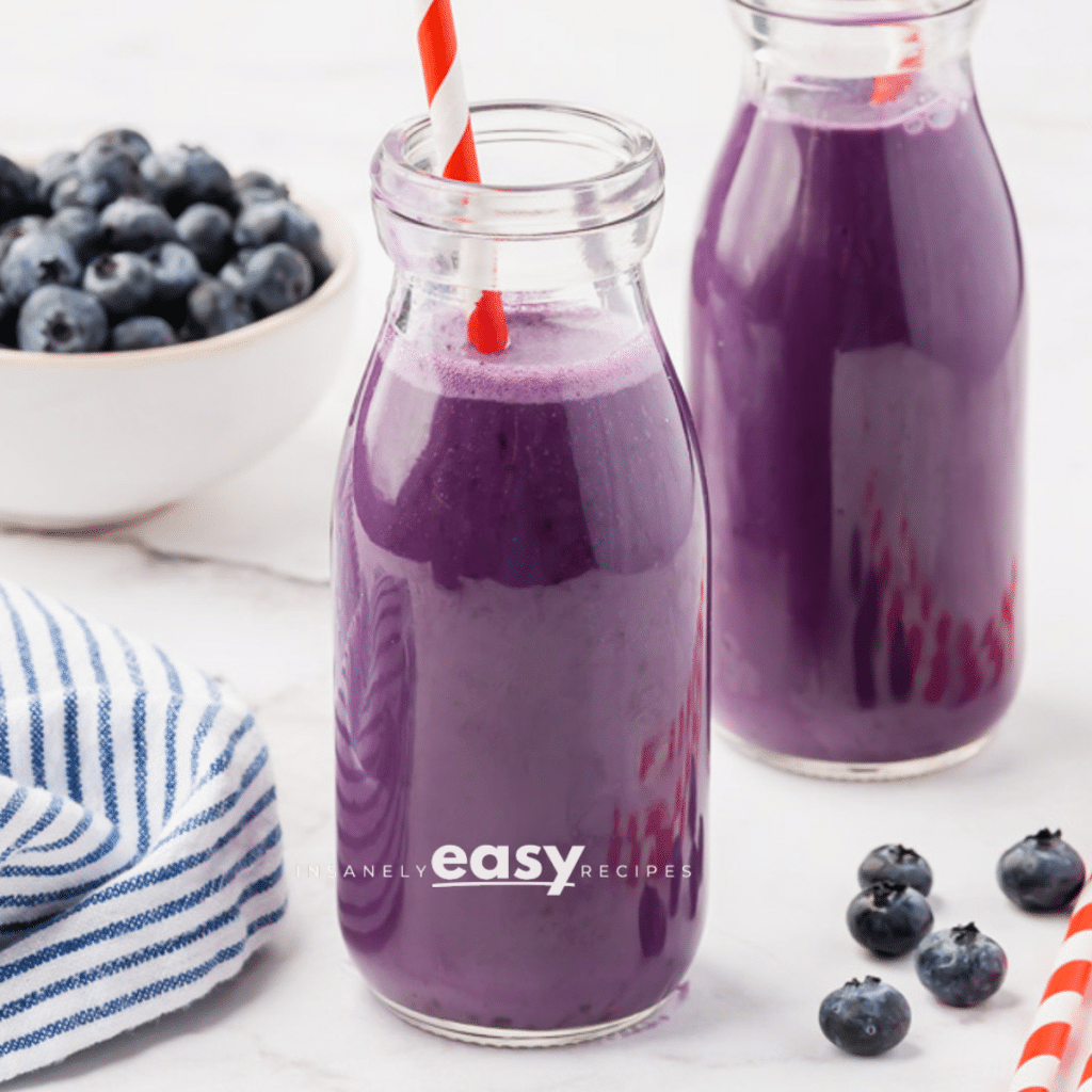 Two small glass jars filled with purple drink. There are red and white straws in each jar, and a bowl of blueberries in the background.