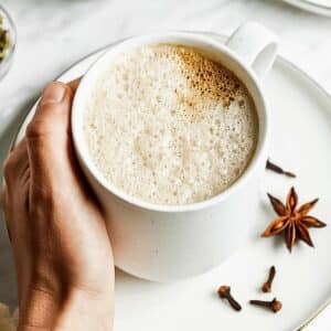 A person holding a white mug filled with a frothy dirty chai latte on a white plate. Next to the mug are a few whole star anise and cloves, adding a touch of spice to the cozy setting.