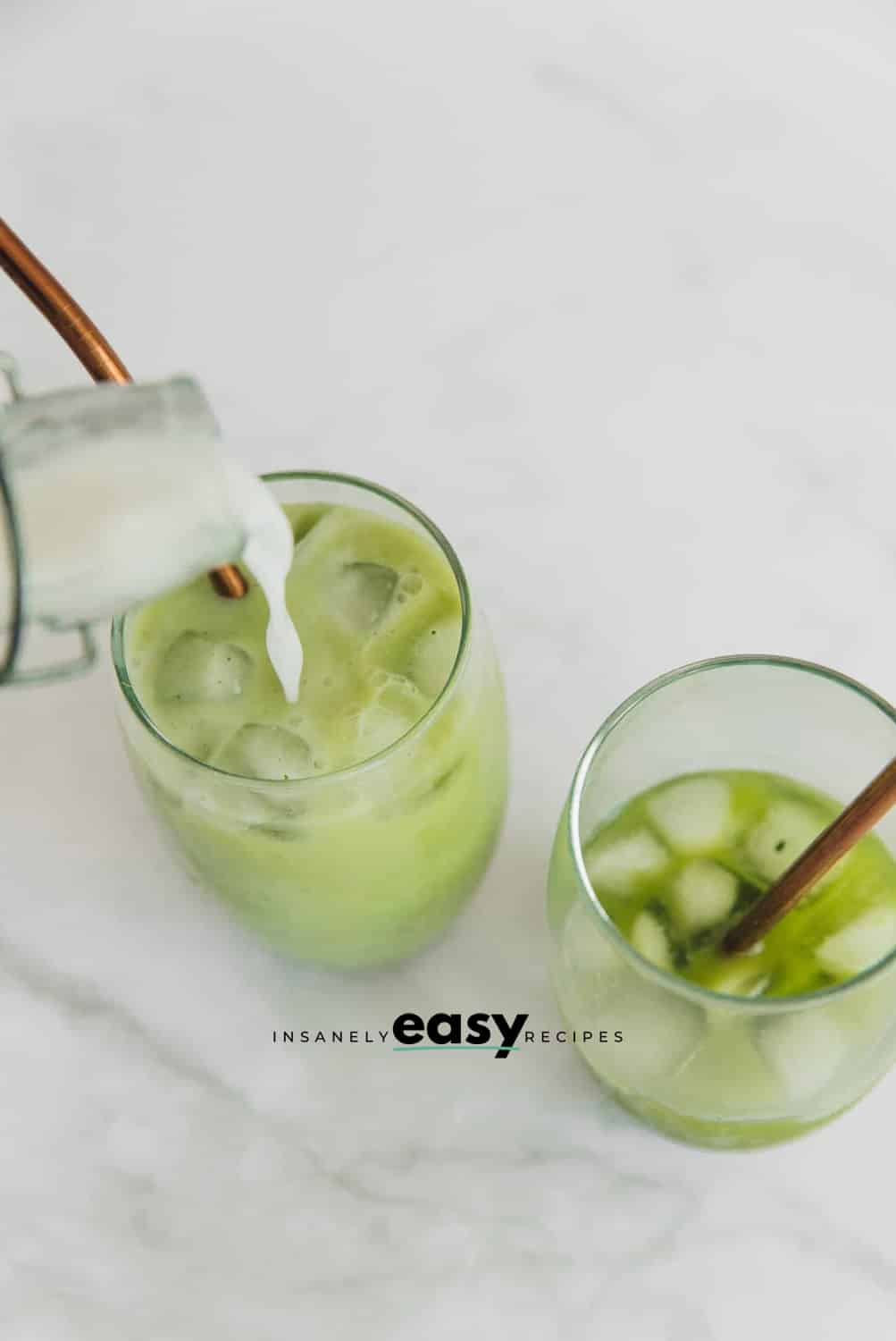 Top view photo of coconut milk being poured into a glass with ice and matcha. The glasses have metal straws.