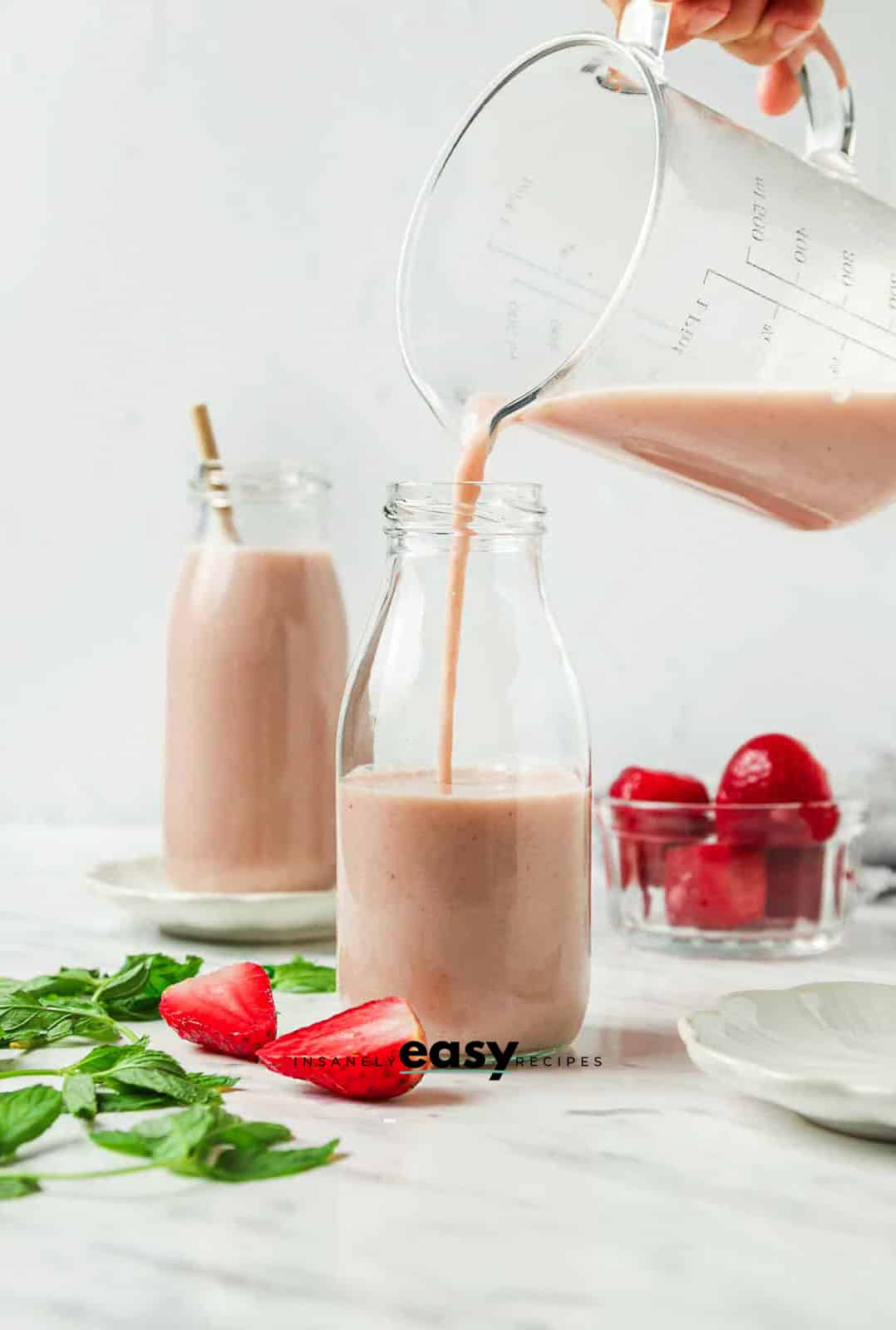 Photo of a hand pouring Strawberry Almond Milk in a tall glass bottle. There is another bottle of Strawberry Almond Milk in the background with a straw and strawberries around the jars.