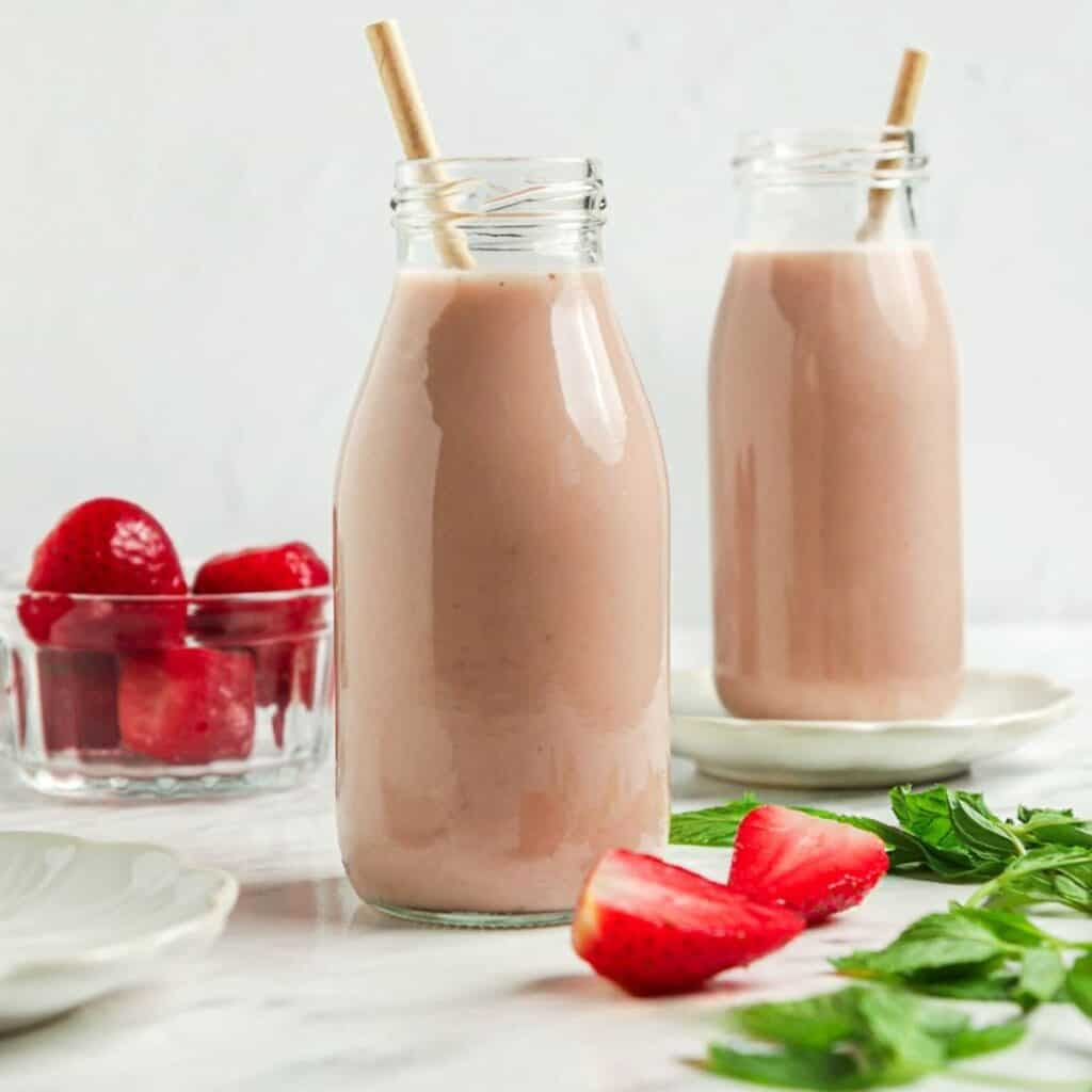 Two glass bottles filled with strawberry almond milk smoothies, each with a straw, are placed on a white surface. A small bowl of whole strawberries and fresh mint leaves are nearby. The background is softly lit, enhancing the creamy pink color of the smoothies.