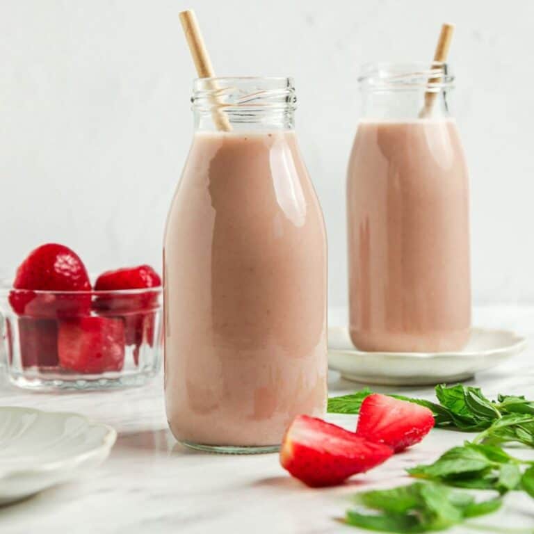 Two glass bottles filled with strawberry almond milk smoothies, each with a straw, are placed on a white surface. A small bowl of whole strawberries and fresh mint leaves are nearby. The background is softly lit, enhancing the creamy pink color of the smoothies.