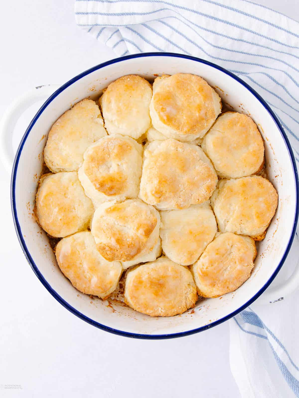 A round baking dish filled with freshly baked golden-brown biscuits. The biscuits are arranged closely together, forming a circular pattern. A striped kitchen towel is partially visible beside the dish on a white surface.