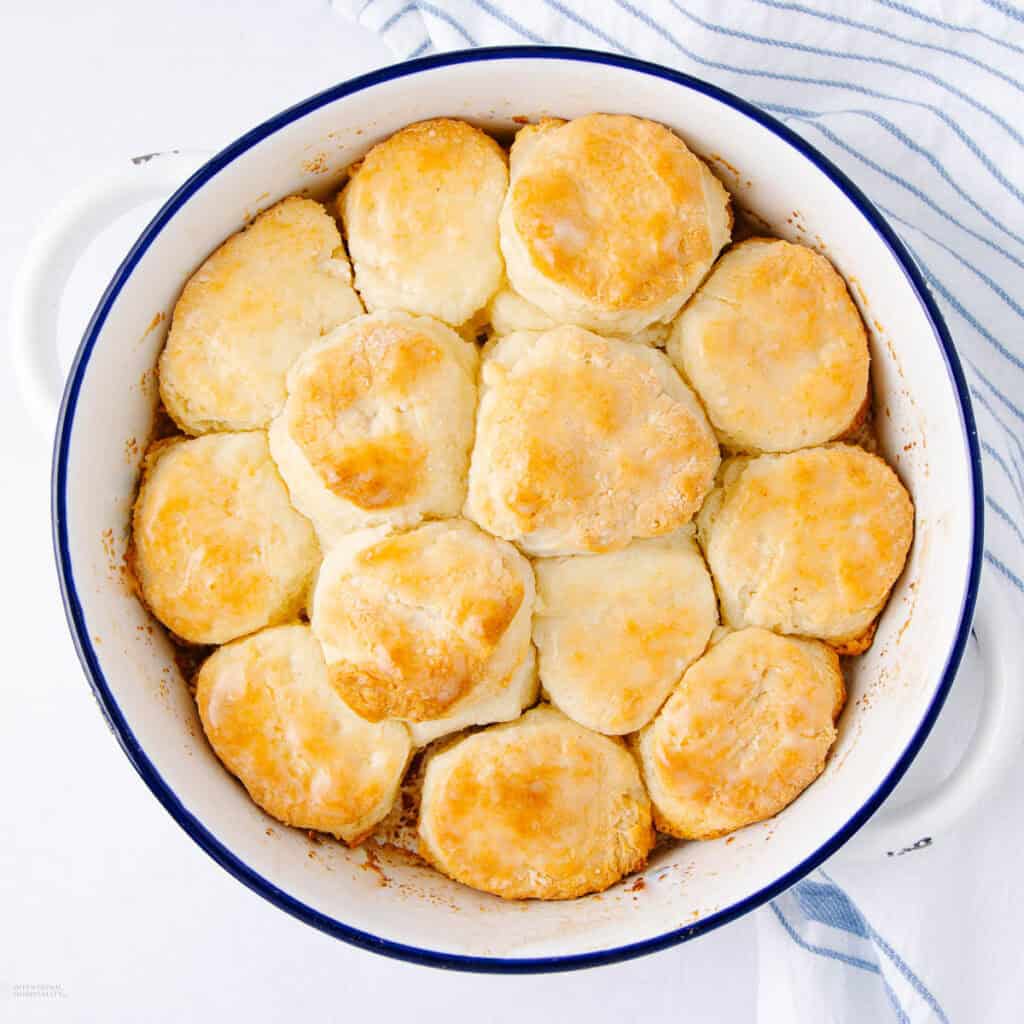 A round baking dish filled with golden brown biscuits arranged in a circular pattern. The biscuits have a crusty top and are slightly touching each other. A striped cloth napkin is partially visible in the background.