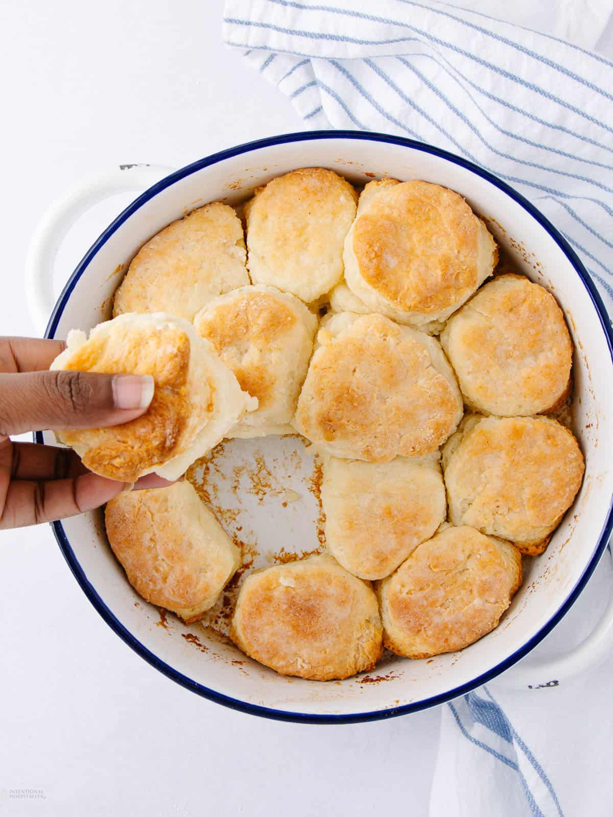 A hand is reaching to pick up a golden-brown biscuit from a round white enamel dish filled with freshly baked biscuits. A striped cloth lies nearby on the white surface.