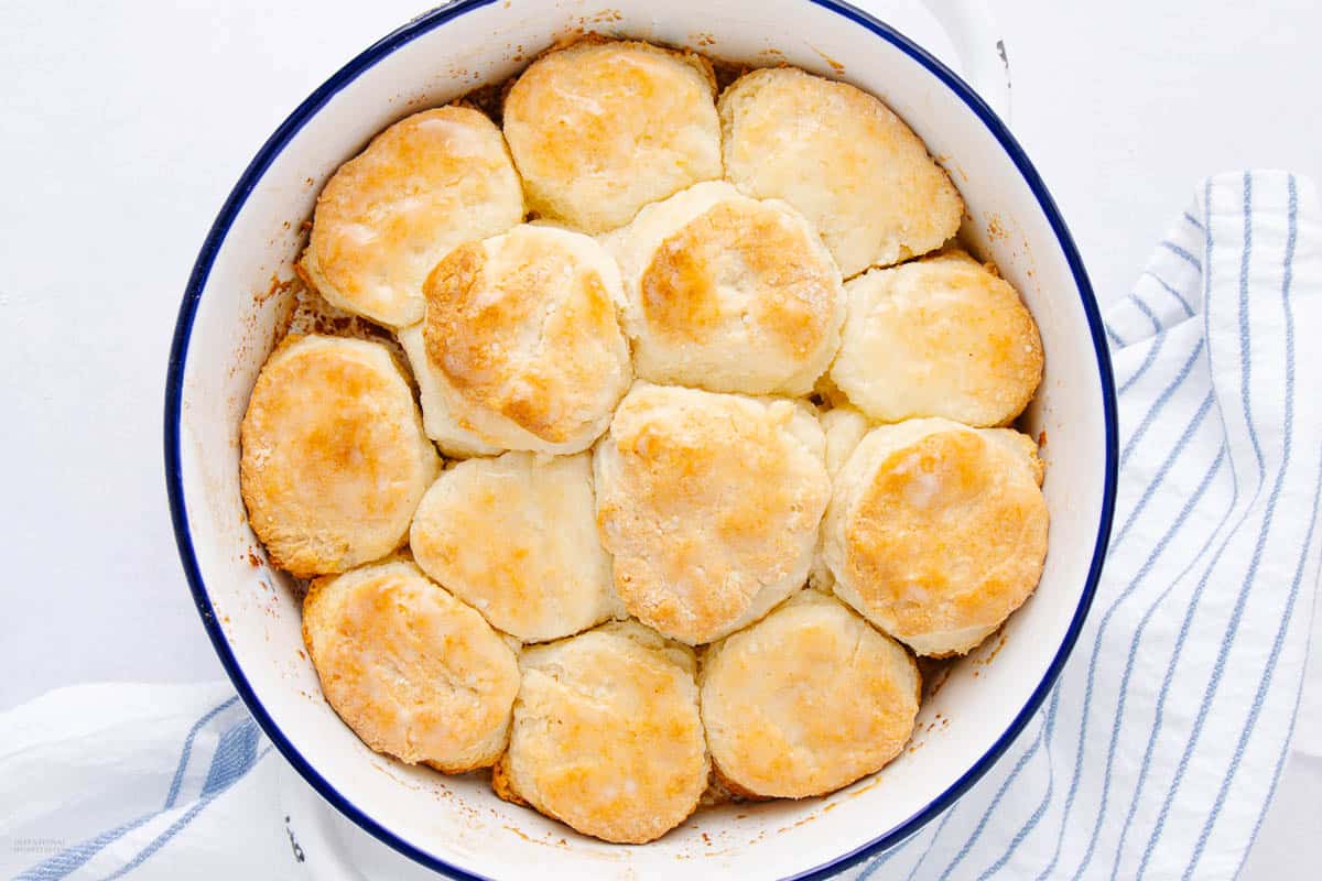 A round white baking dish filled with golden-brown baked biscuits arranged neatly. A striped kitchen towel is partially visible beside the dish on a light background.