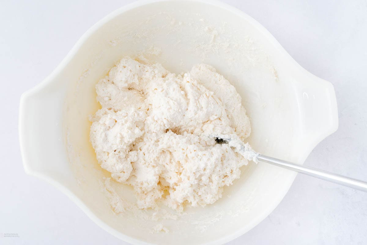 A white mixing bowl filled with a thick, creamy dough or batter. A metal spoon is partially submerged in the mixture. The bowl rests on a light-colored surface.