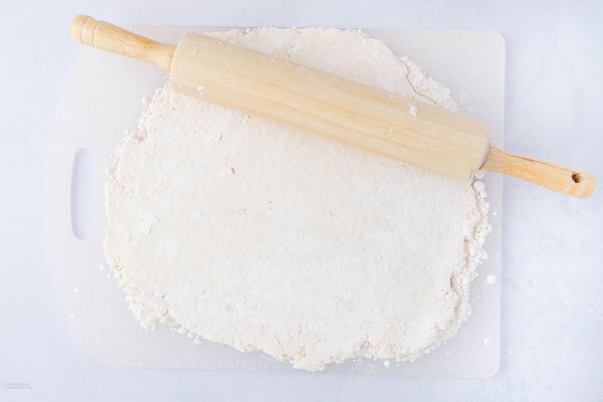 A wooden rolling pin resting on flattened, floured dough on a white surface.