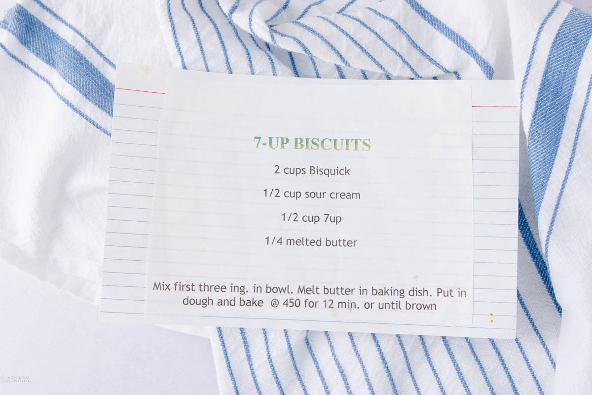 A handwritten recipe for 7-Up Biscuits on lined paper, placed on a white cloth with blue stripes. Ingredients include Bisquick, sour cream, 7-Up, and melted butter. Baking instructions are also listed.