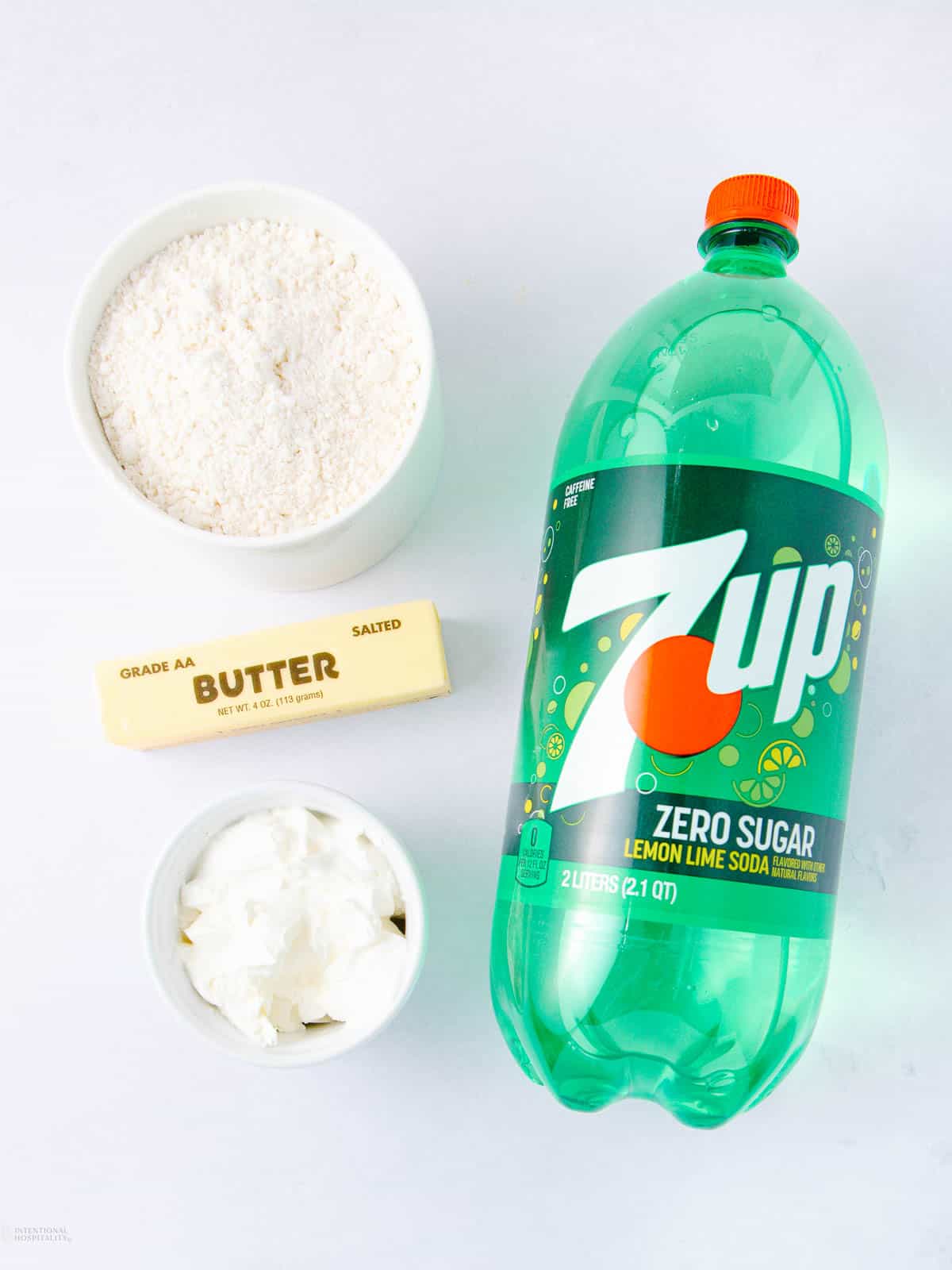 A 2-liter bottle of 7UP Zero Sugar is placed next to a stick of salted butter, a bowl of white powder (likely flour), and a bowl of white solid ingredient (possibly shortening) on a white surface.