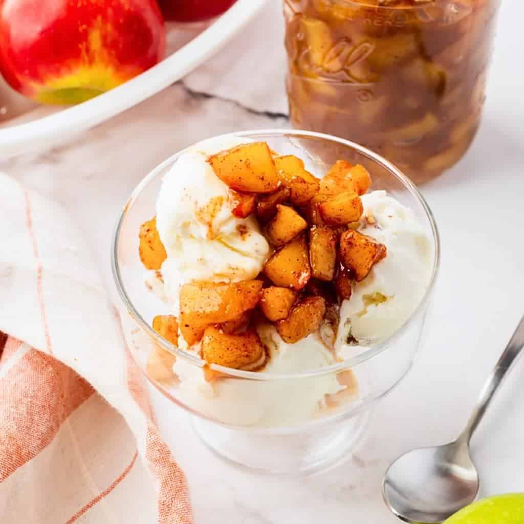 A glass dish filled with vanilla ice cream topped with cinnamon-spiced apple chunks. A jar of apple topping and fresh apples are in the background, alongside a striped napkin and a spoon on a marble countertop.