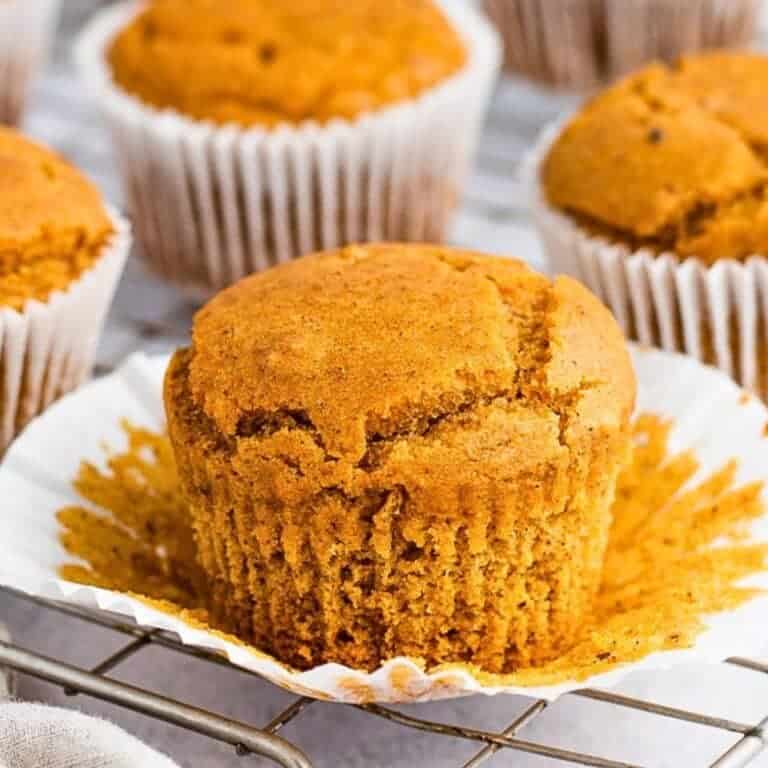 A freshly baked pumpkin muffin sits in an open white paper liner on a cooling rack. Several similar muffins are visible in the background, each with a golden-brown, slightly cracked top.