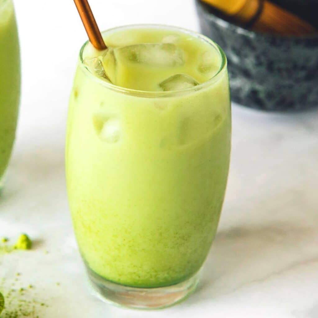 A glass of iced matcha latte with a metal straw on a light surface. The creamy green beverage has ice cubes floating on top and a bowl with matcha powder is blurred in the background.