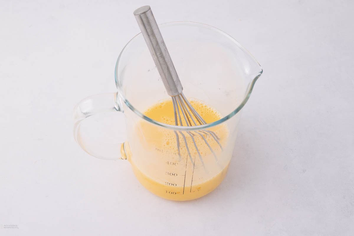 A clear glass measuring cup filled with beaten eggs, placed on a white surface. A metal whisk rests inside the cup, with measurement markings visible on the side of the glass.