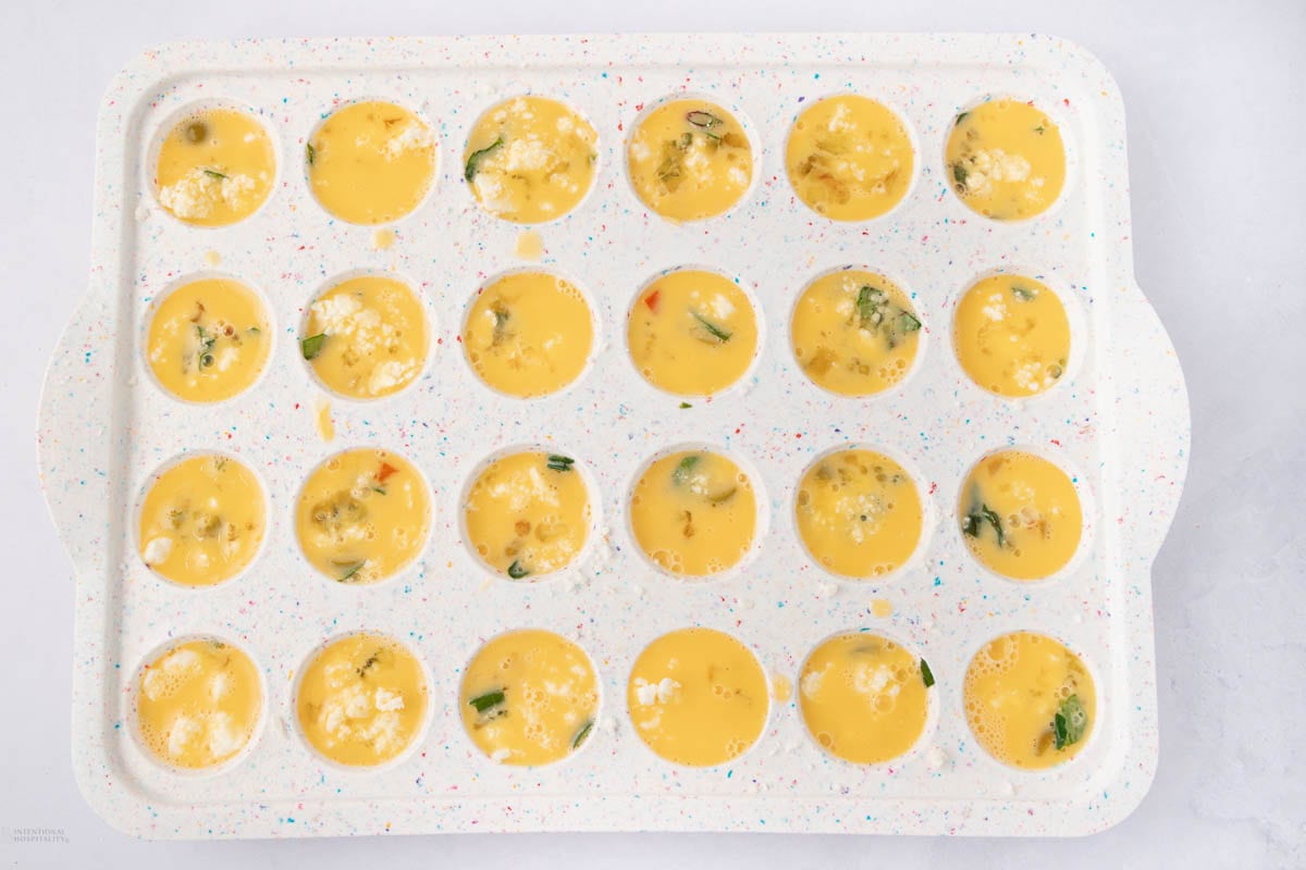 A white, speckled silicone mold filled with raw beaten eggs combined with vegetables and cheese, ready to be cooked. The mold has two handles, and each cavity contains a portion of egg mixture.