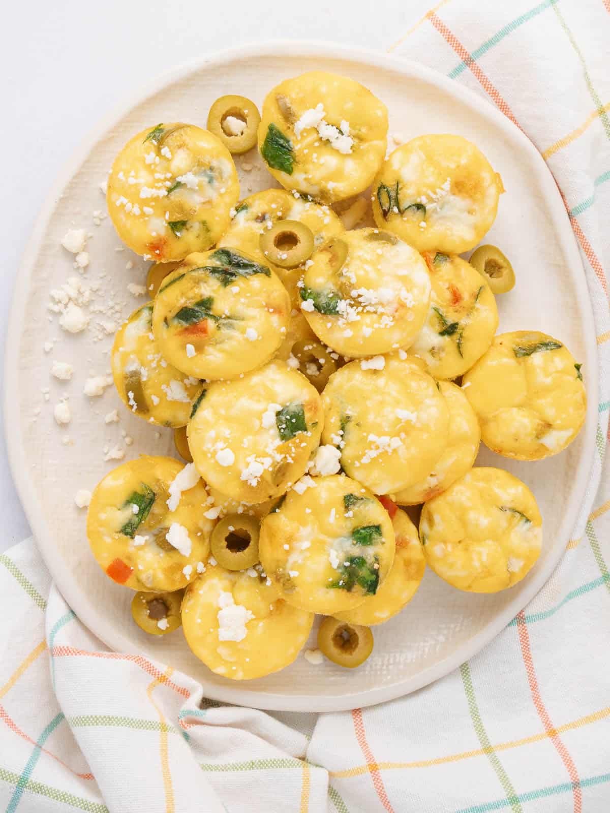 A plate of mini egg muffins garnished with crumbled cheese and sliced olives. The muffins are mixed with spinach and red bell peppers. The plate is on a colorful plaid napkin.