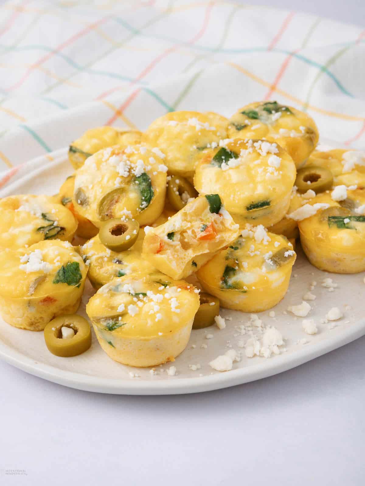 A plate of mini egg muffins topped with crumbled cheese, featuring visible slices of green olives and spinach. One muffin is cut in half, highlighting the filling. The background includes a white cloth with a colorful grid pattern.