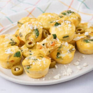 A plate of mini egg muffins with vegetables and olives, topped with crumbled feta cheese. The muffins are golden brown and placed against a checkered cloth background.