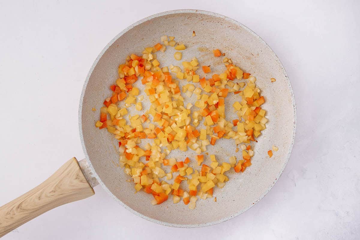 A frying pan with a light wooden handle contains diced orange and yellow bell peppers and onions being sautéed. The ingredients are spread evenly, showing a mix of vibrant colors against the light background of the pan.