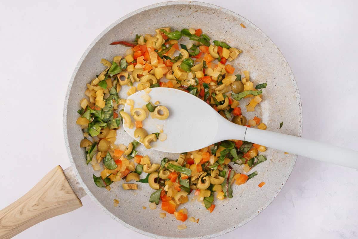 A frying pan containing a mix of chopped vegetables, including yellow bell peppers, red peppers, spinach, and sliced green olives. A white spatula rests on top, and the pan has a wooden handle. The background is a white surface.