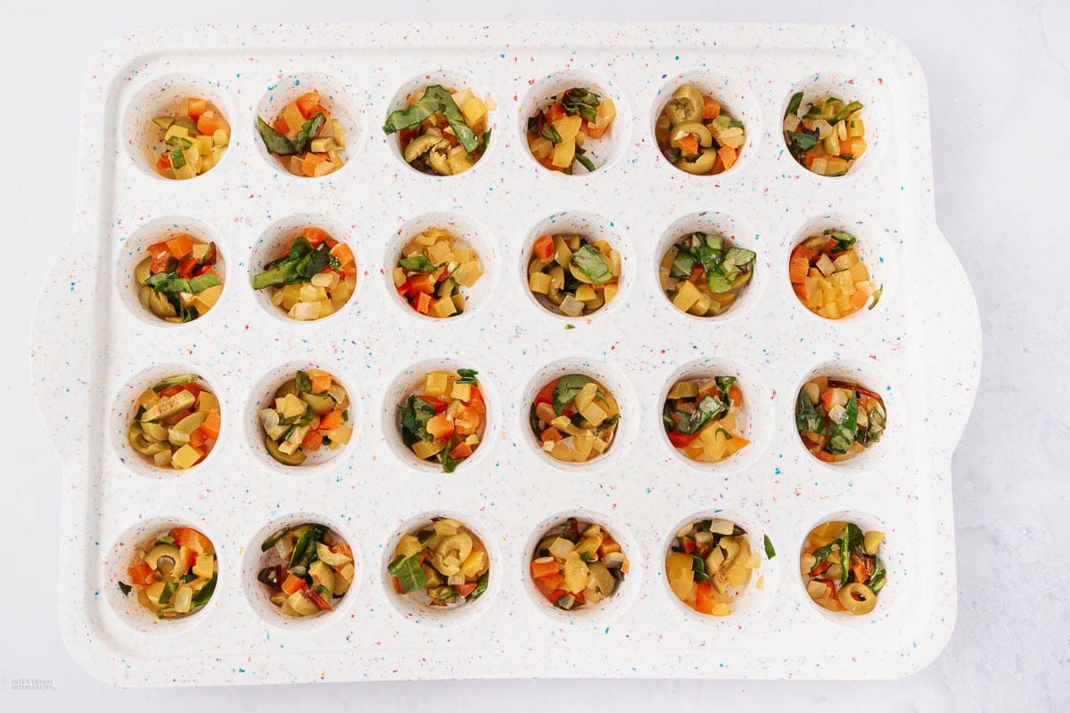 A muffin tray filled with chopped vegetables, including green olives, carrots, and herbs, evenly distributed in each cup. The tray is white with colorful specks, and it is placed on a light surface.