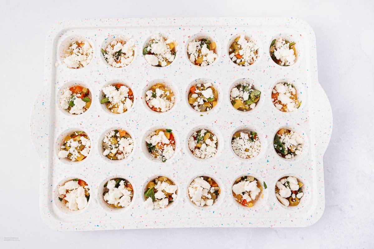 A white muffin tray filled with 24 mini quiches, topped with crumbled feta cheese, diced vegetables, and herbs. The tray is set on a light surface.