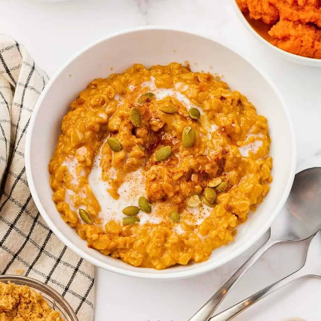 A bowl of creamy pumpkin oatmeal topped with pumpkin seeds and a sprinkle of cinnamon. A checkered cloth and a spoon are placed beside the bowl, with an additional bowl of pumpkin puree partially visible.