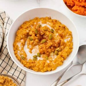 A bowl of creamy pumpkin oatmeal topped with pumpkin seeds and a sprinkle of cinnamon. A checkered cloth and a spoon are placed beside the bowl, with an additional bowl of pumpkin puree partially visible.