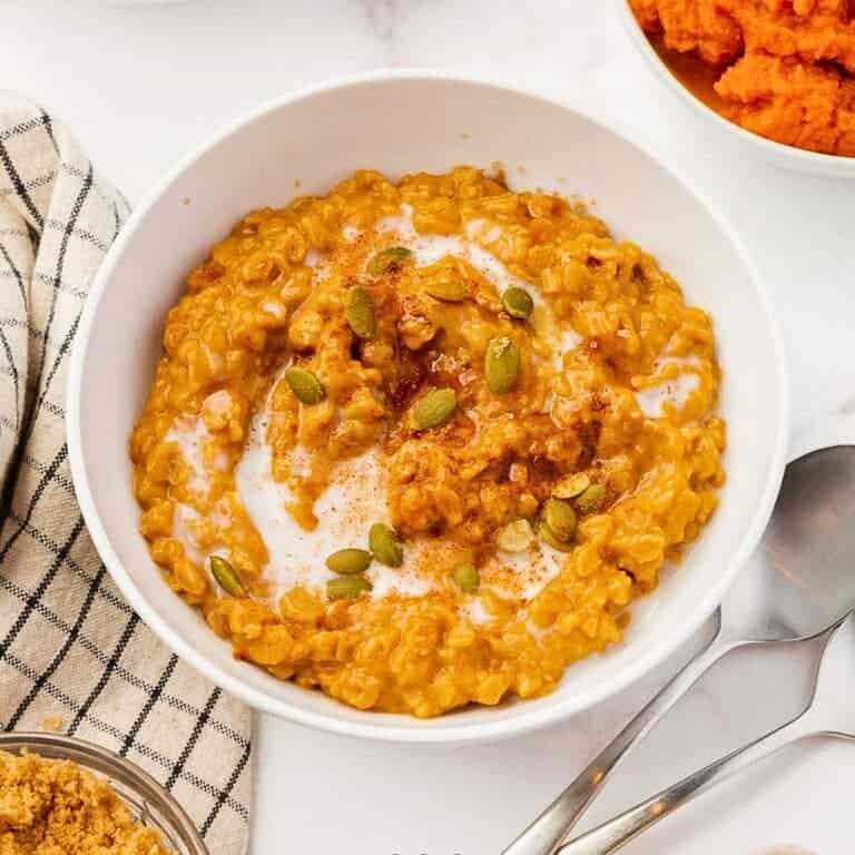 A bowl of creamy pumpkin oatmeal topped with pumpkin seeds and a sprinkle of cinnamon. A checkered cloth and a spoon are placed beside the bowl, with an additional bowl of pumpkin puree partially visible.