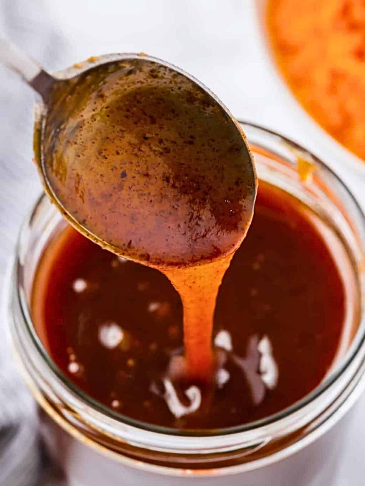 A close-up of a spoon pouring thick, reddish-brown sauce back into a jar. The sauce has a glossy texture with visible seasoning specks. A blurred orange object is in the background.