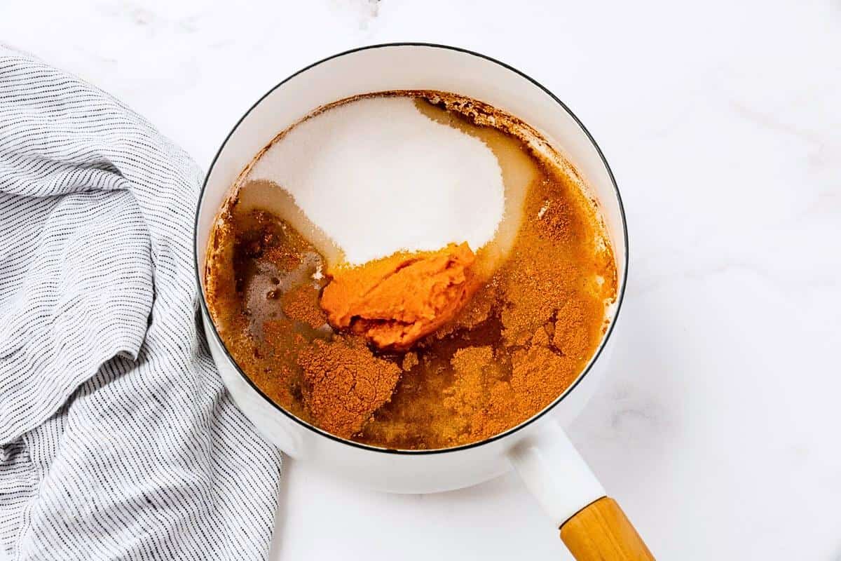 A saucepan on a white surface contains ingredients like pureed pumpkin, sugar, and spices, ready to be mixed. A striped cloth is placed nearby.