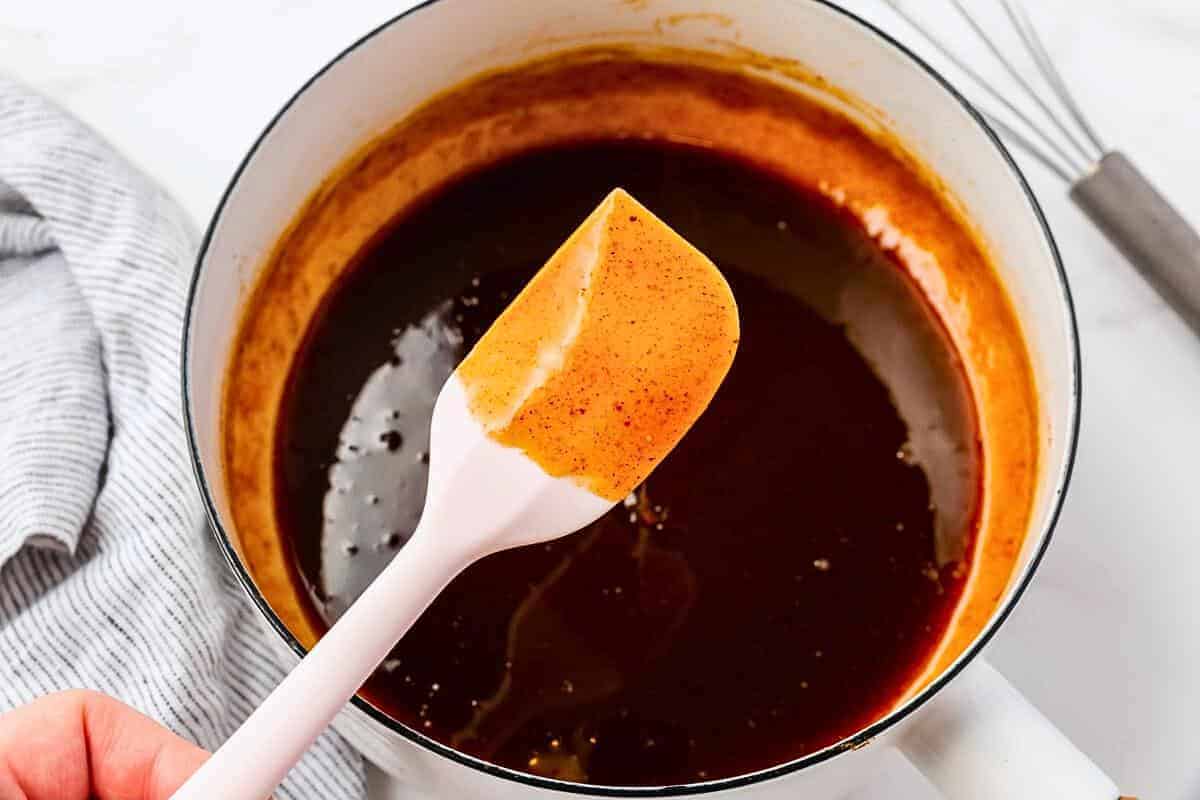 A close-up of a pot filled with smooth, dark brown sauce. A spatula is held above the pot, showing the sauce’s glossy texture. A whisk is partially visible on the right, and a striped cloth is on the left.