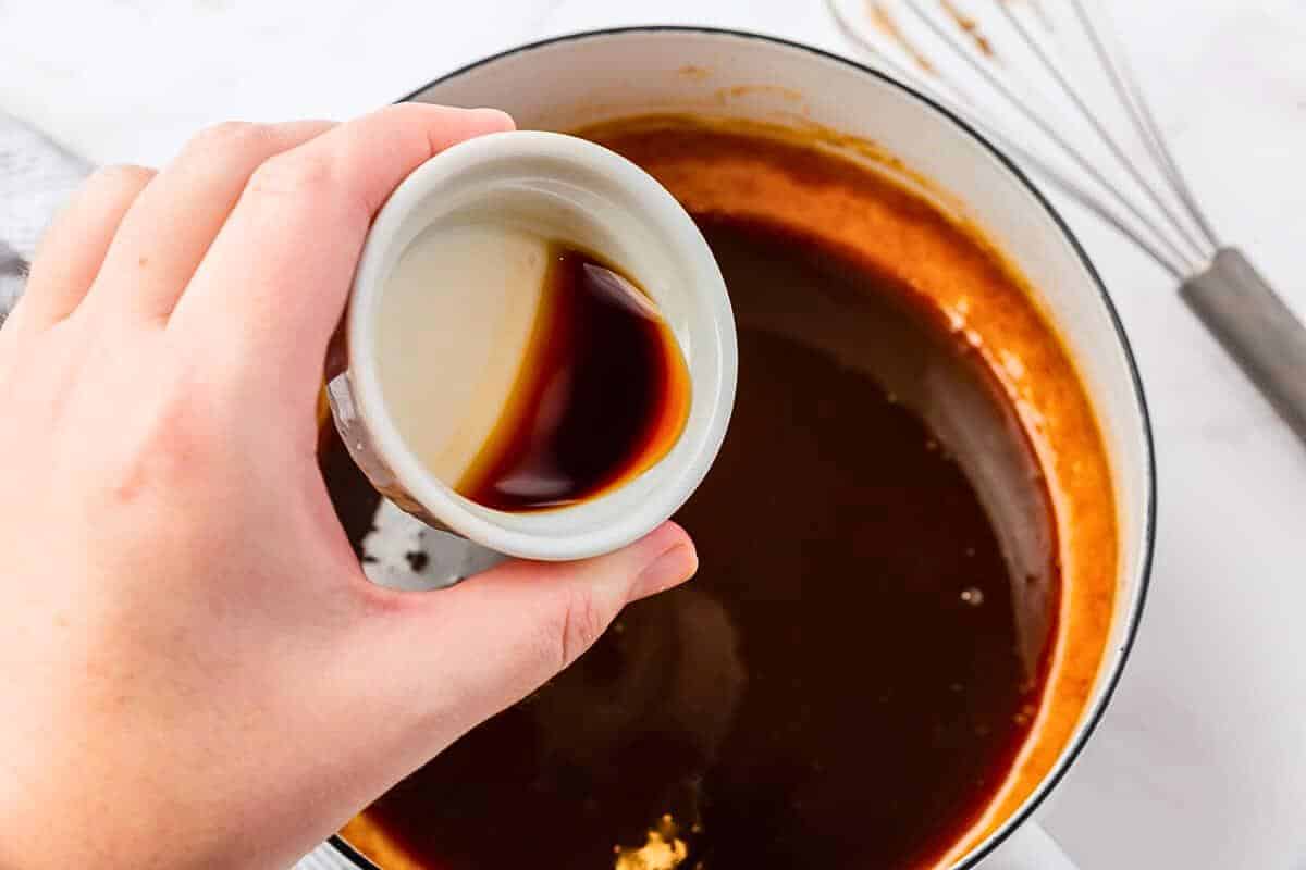 A hand pouring soy sauce from a small white cup into a pot containing a dark, simmering liquid. A whisk is on the countertop nearby.